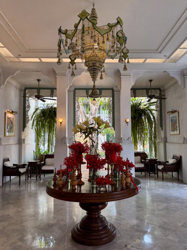 Elegant lobby of The Inside House hotel in Chiang Mai, Thailand, featuring a traditional Thai chandelier, marble floors, lush greenery, and a centerpiece table adorned with vibrant red floral arrangements.
