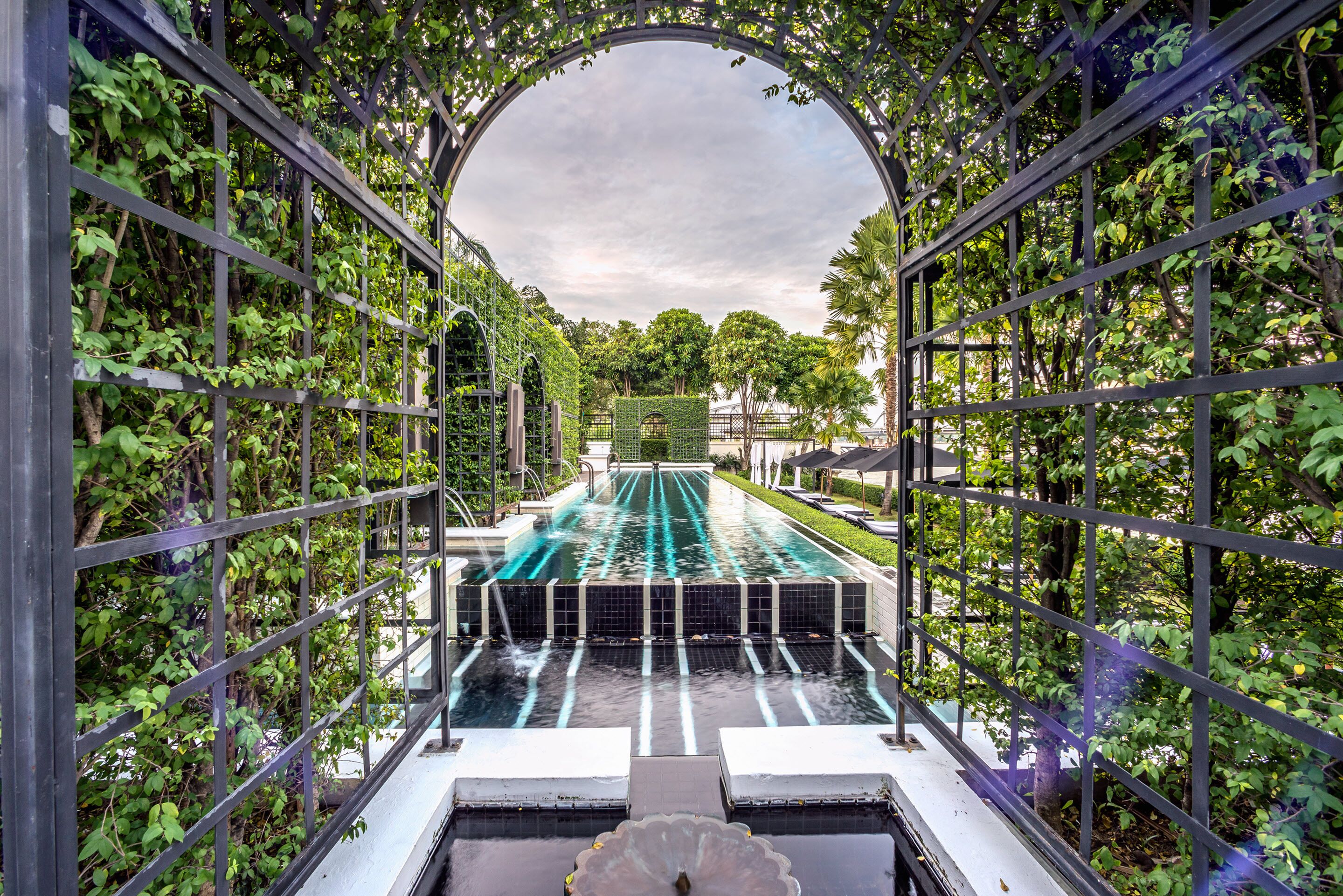 Serene view of The Siam Hotel’s elegant lap pool framed by lush green trellises, with symmetrical lines, cascading fountains, and a tranquil garden backdrop.