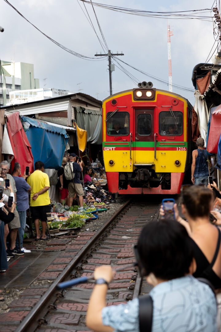 A brightly colored train slowly approaches through the narrow Maeklong Railway Market in Thailand as tourists and vendors press to the edges, capturing the unique moment with their phones.