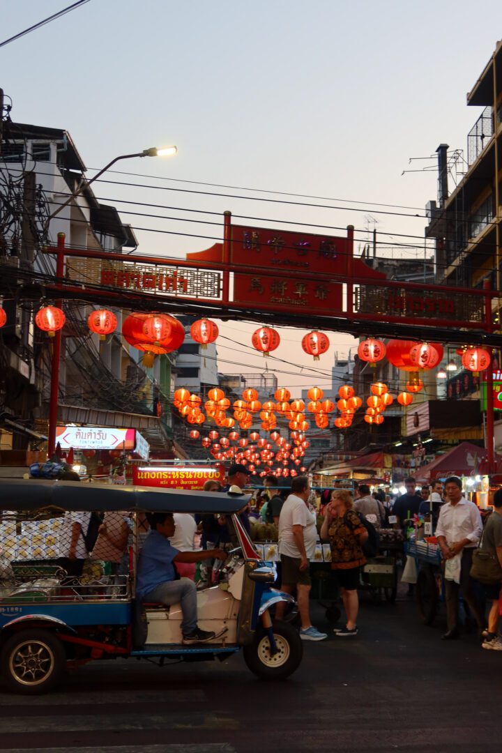 A tuk-tuk weaves through the bustling streets of Bangkok’s Chinatown at dusk, where red lanterns hang overhead and food stalls fill the night with vibrant energy.