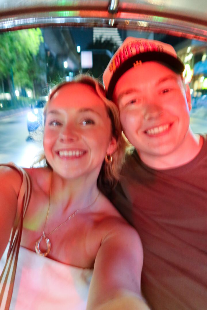 Joyful couple taking a blurry selfie during a nighttime tuk-tuk ride through the glowing city streets of Bangkok.
