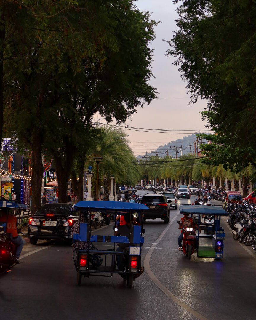 Tuk-tuks and motorbikes driving along a busy tree-lined street in Ao Nang, Krabi, with shops and traffic during the golden hour.