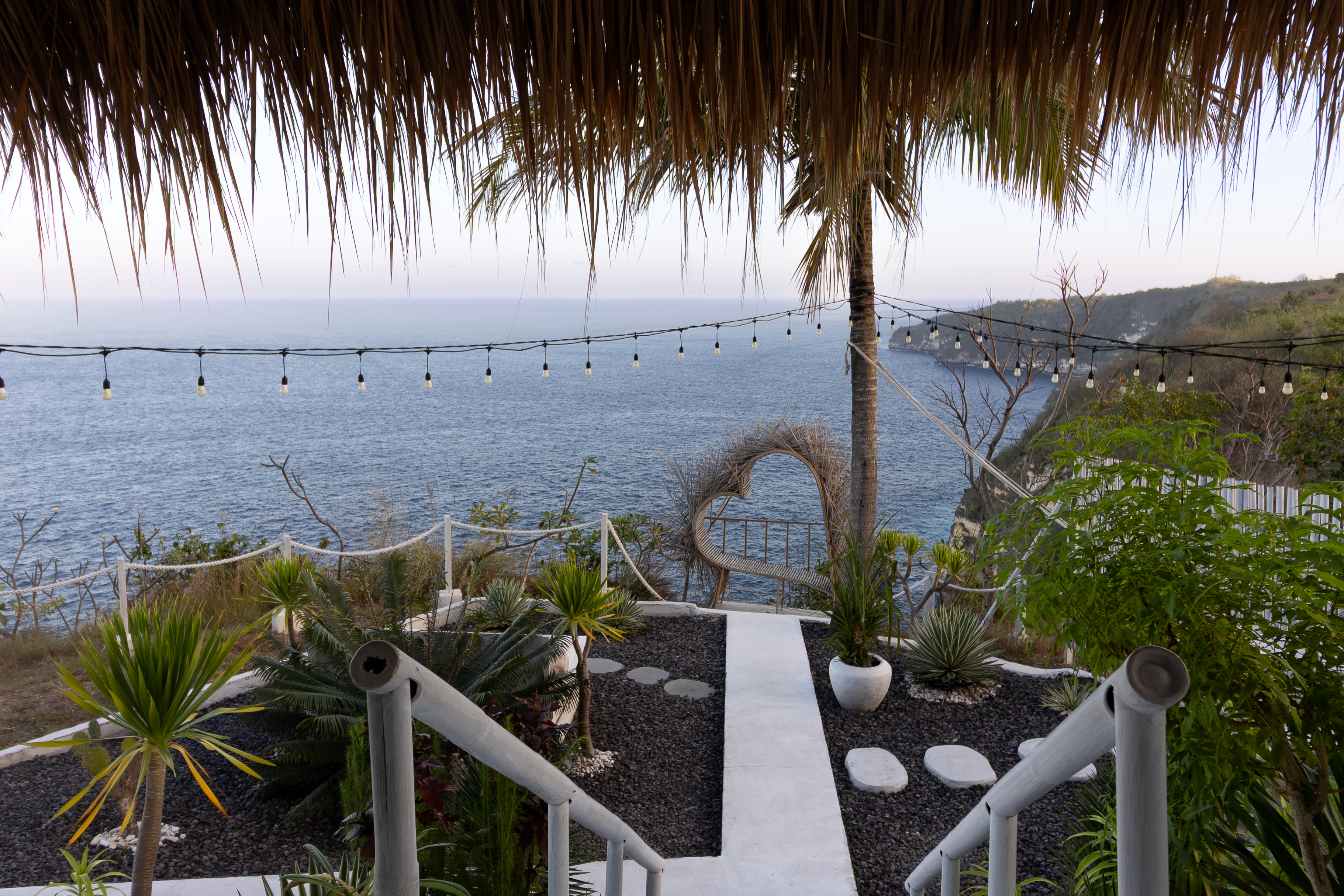 Cliffside view from Tropical Glamping Nusa Penida featuring a heart-shaped bamboo photo spot, string lights, and a sweeping panorama of the ocean at dusk.