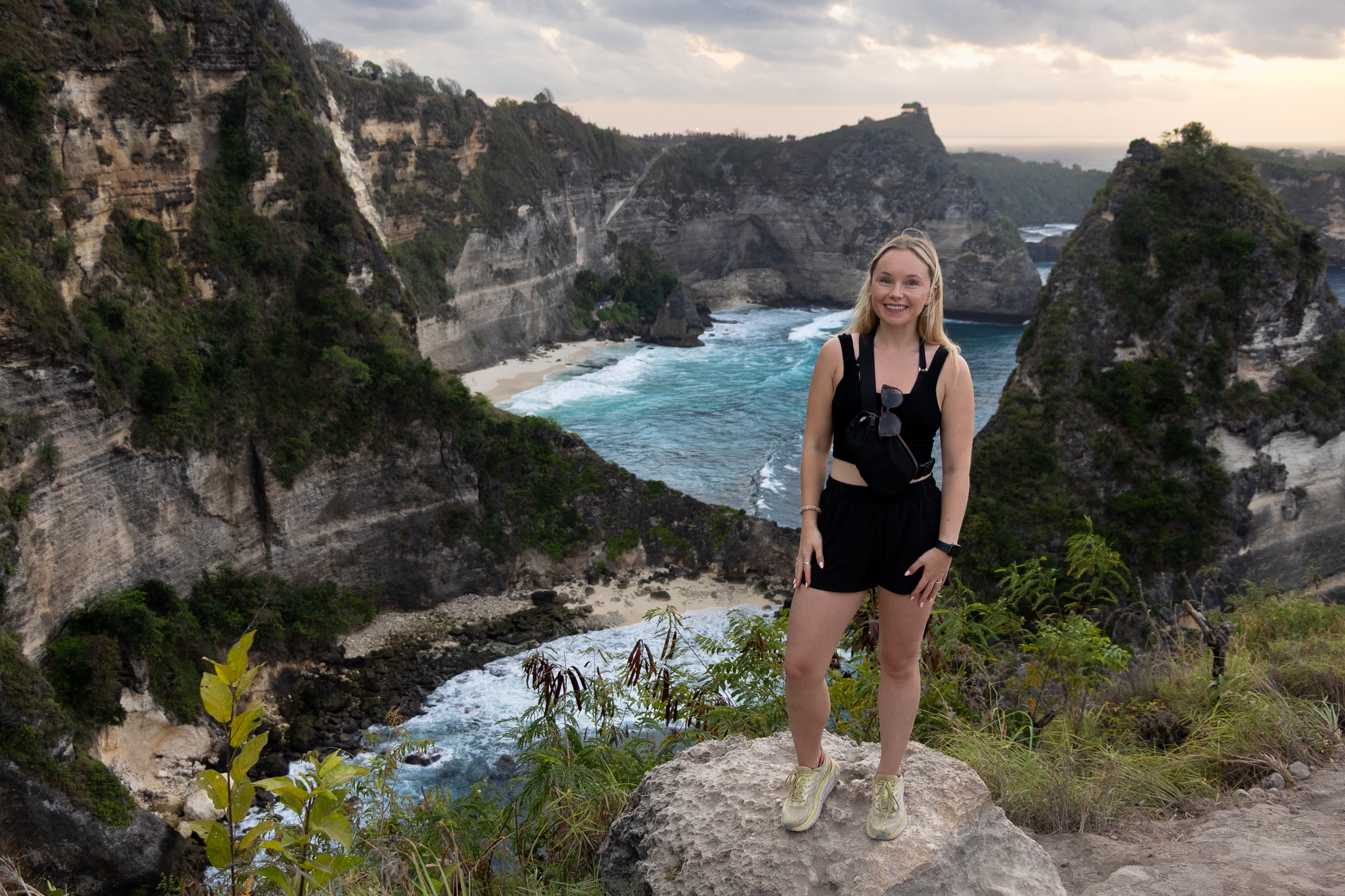 Woman standing on a cliffside rock at Thousand Islands Viewpoint in Nusa Penida, overlooking dramatic sea stacks and turquoise waves below.