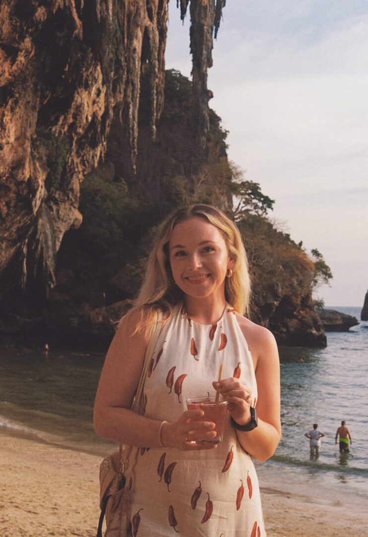 Woman holding a cocktail and smiling on Railay Beach at sunset, with towering limestone cliffs and calm sea in the background.