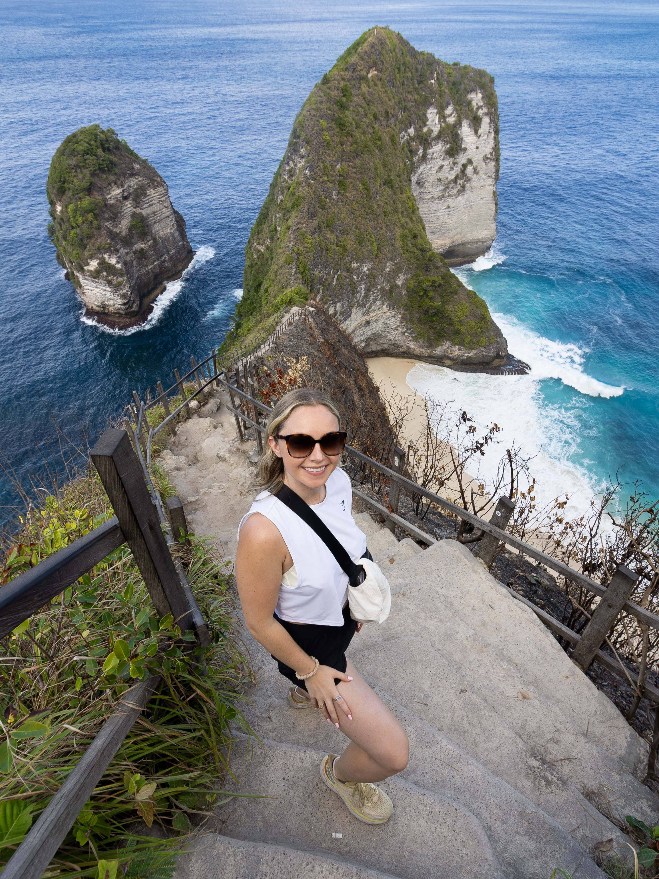 Smiling woman posing on the famous cliffside stairs at Kelingking Beach, with the dramatic T-Rex-shaped rock formation and turquoise sea behind her.