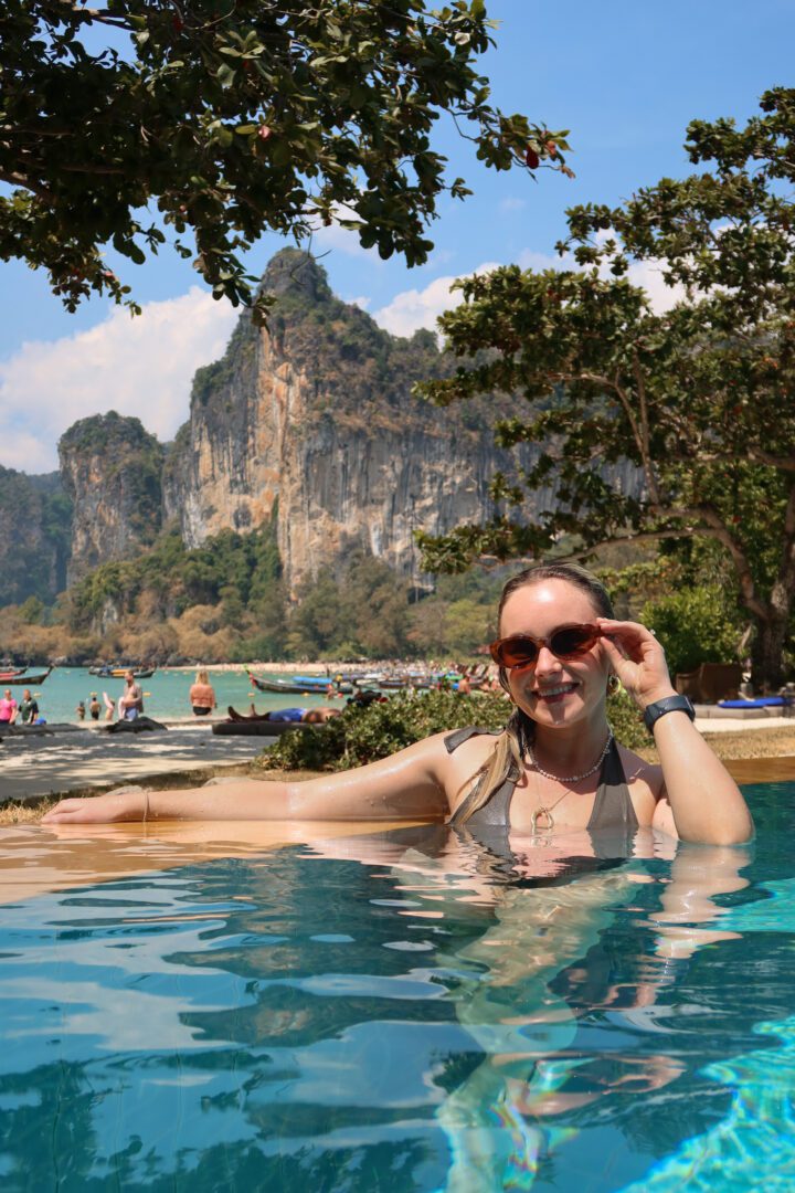 Woman relaxing in the infinity pool at Rayavadee Resort in Railay, gazing out at the beach and surrounding cliffs under a sunny blue sky.