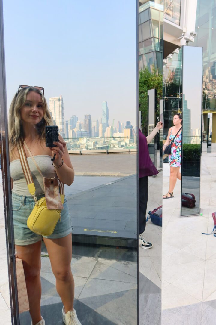 A woman captures a mirror selfie on the rooftop of ICONSIAM shopping mall, with the Bangkok skyline—including the iconic MahaNakhon tower—visible in the background.
