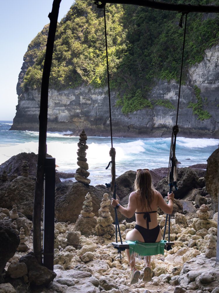 Woman in a swimsuit sitting on a beach swing facing the crashing waves and stacked rock cairns of Tembeling Beach.
