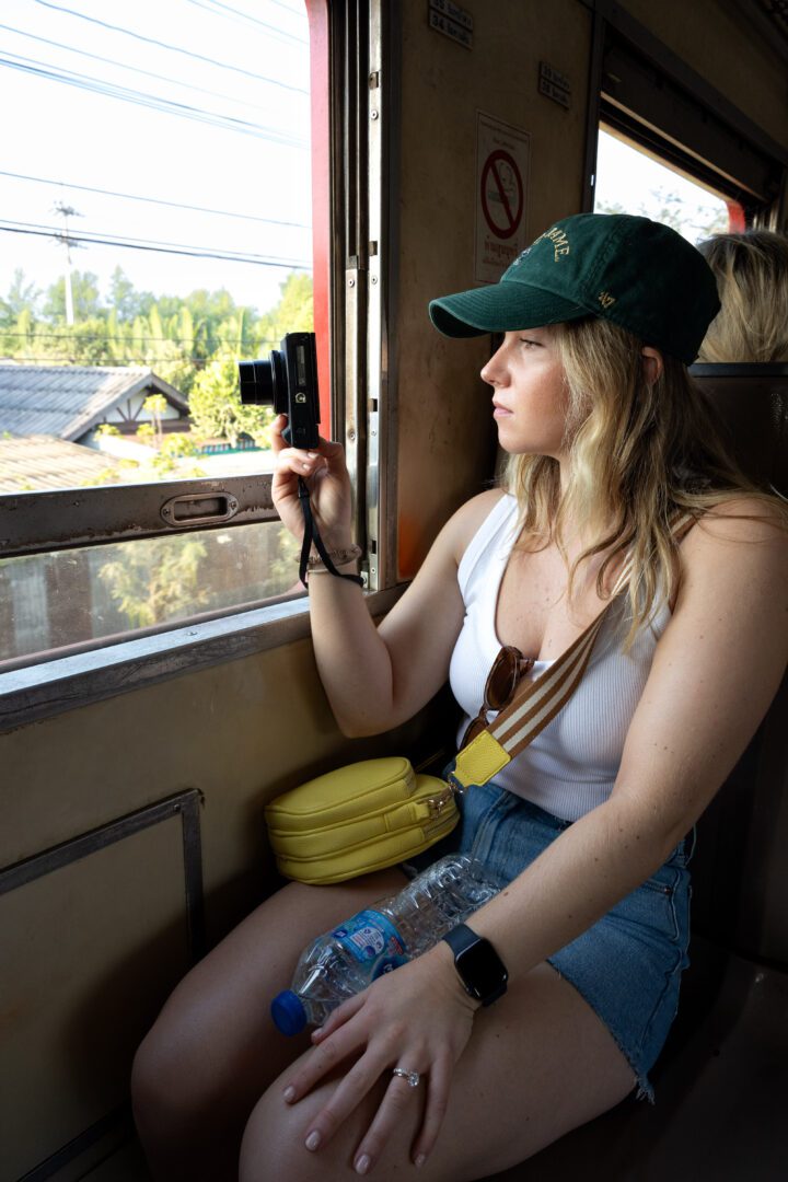 A woman rides the train to Maeklong Railway Market, capturing the passing scenery through the open window with her camera on a sunny day in Thailand.