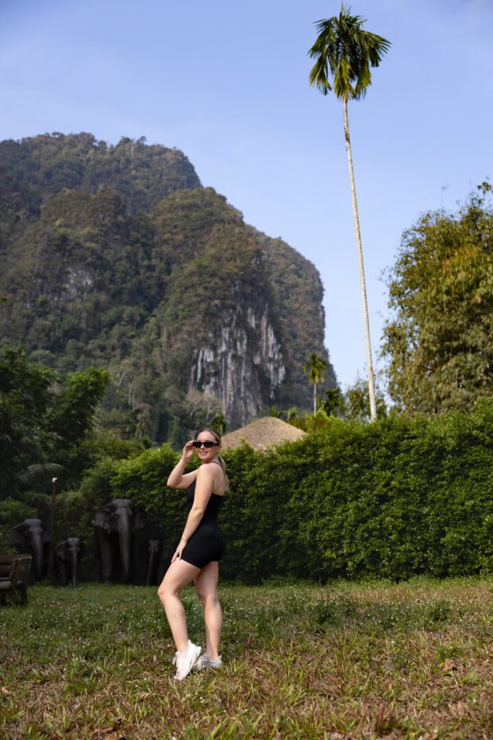 A woman poses confidently on a grassy field at Elephant Hills Jungle Camp, with limestone cliffs and lush greenery rising behind her.