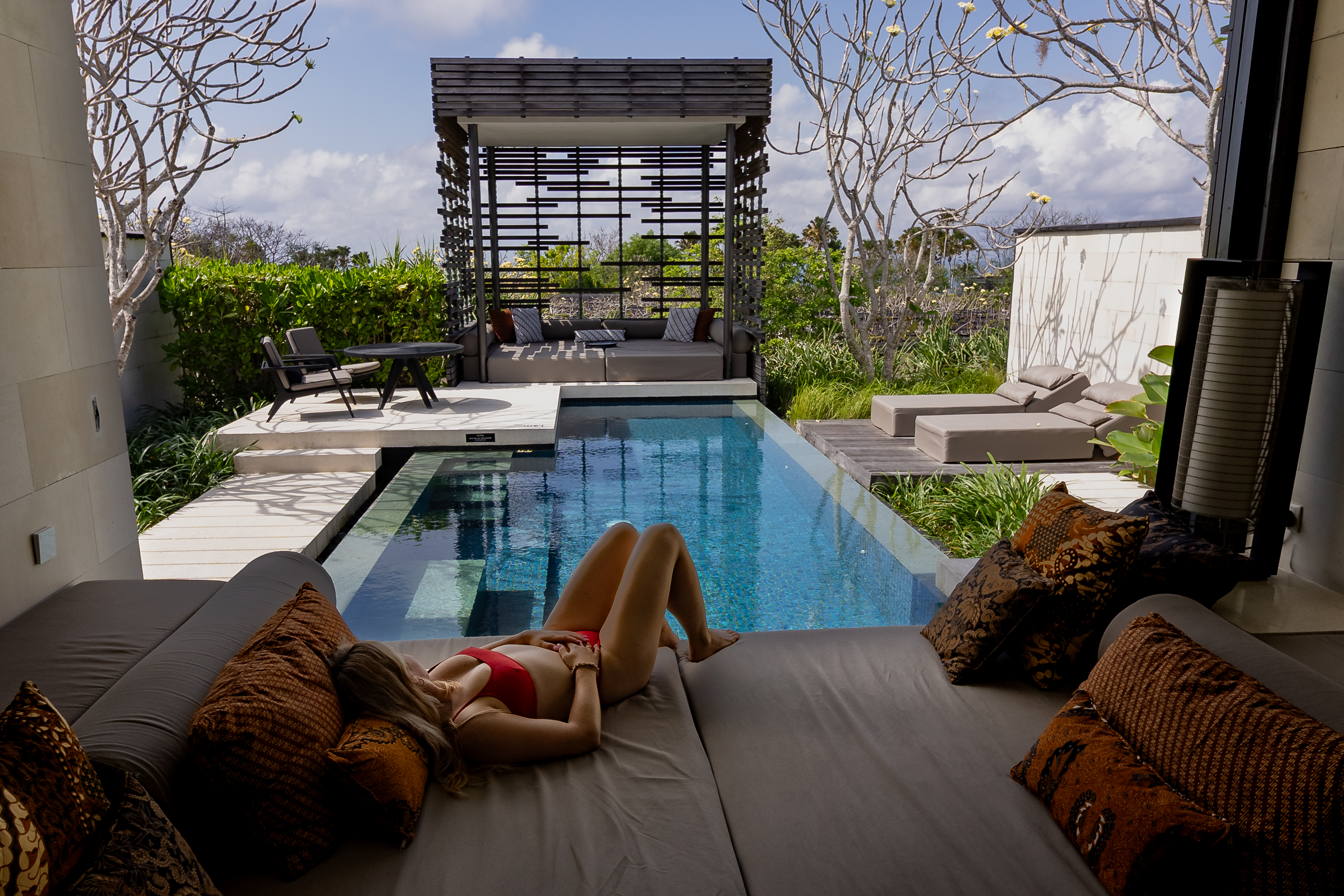 Woman in a red bikini reclining on a daybed beside a private pool at Alila Villas Uluwatu, framed by tropical landscaping and elegant outdoor furnishings.