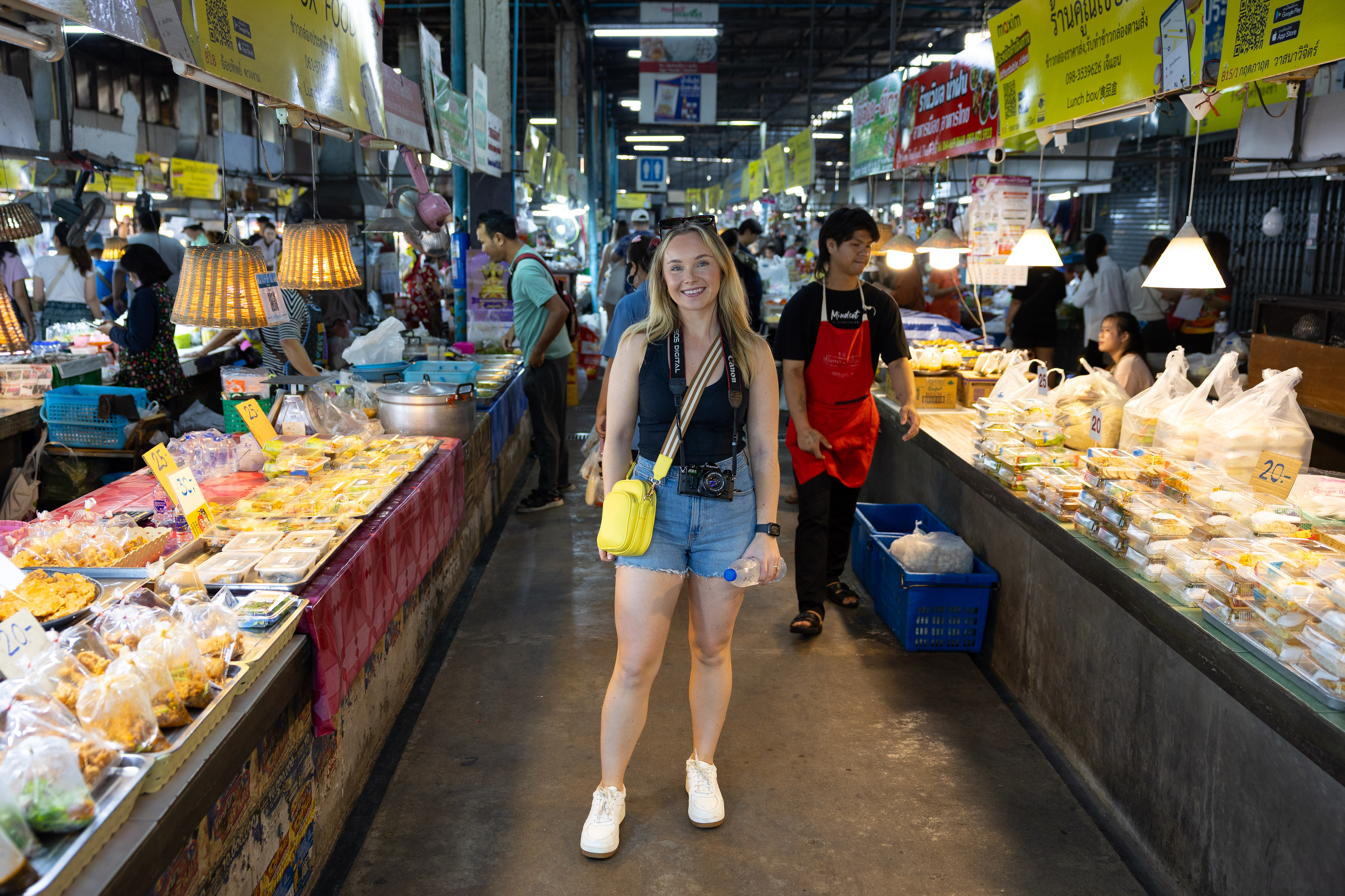 Smiling woman exploring a bustling local market in Chiang Mai, Thailand, surrounded by food vendors selling traditional Thai snacks, packaged meals, and fresh ingredients under bright hanging lights.