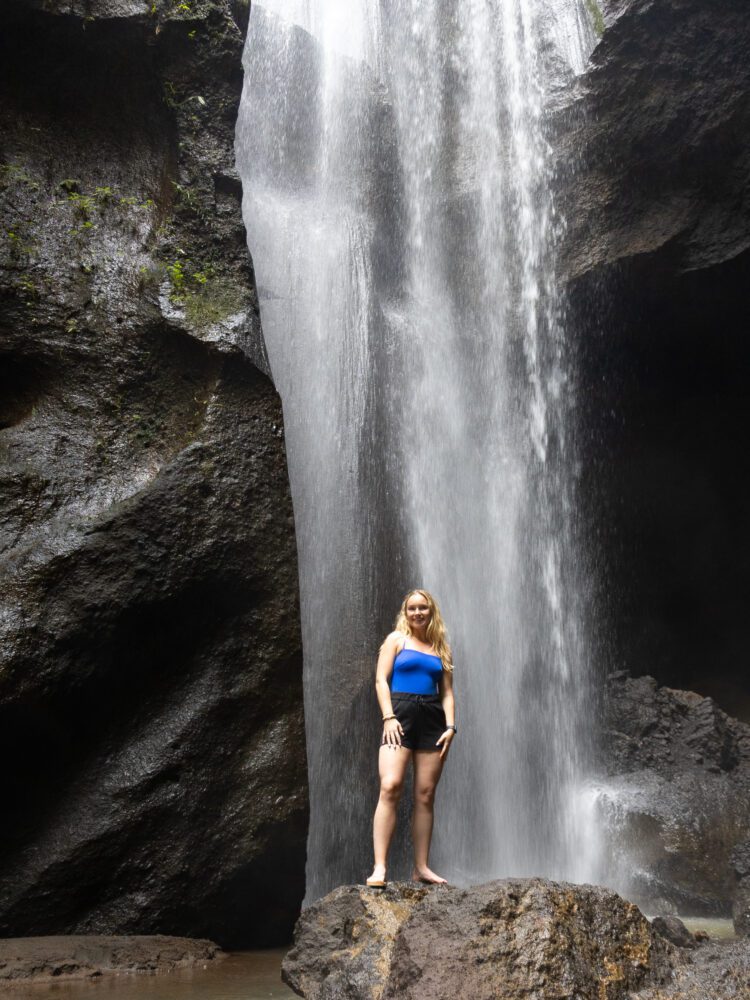 Woman standing confidently on a rock beneath a dramatic waterfall inside a cave in Bali.