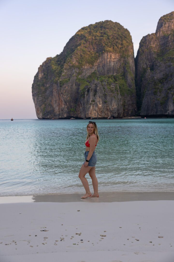 Woman posing on the soft white sand of Maya Bay, Koh Phi Phi, with calm aqua water and dramatic cliffs in the pastel morning glow.