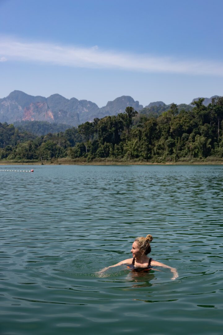 A woman enjoys a refreshing swim in the clear green waters of Cheow Lan Lake, with jungle-covered mountains forming a breathtaking backdrop at Elephant Hills Lake Camp.