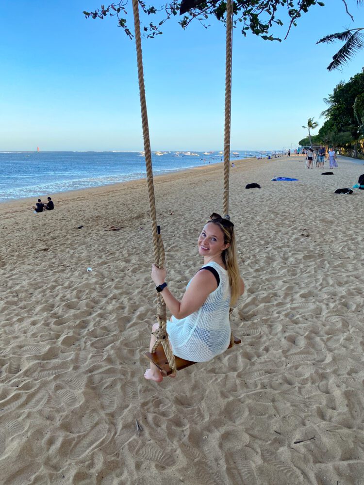 Woman smiling on a wooden beach swing under tree branches at Sanur Beach during sunset.