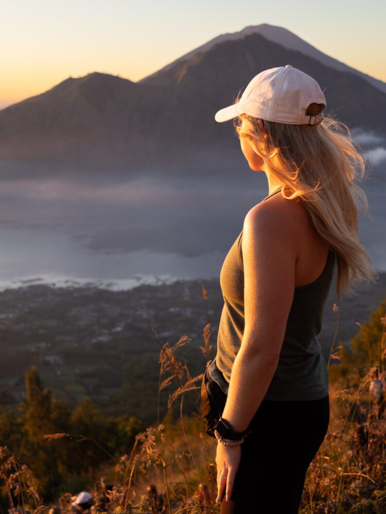 Woman standing on a hillside at sunrise, overlooking Mount Batur and Lake Batur after a sunrise trek in Bali.