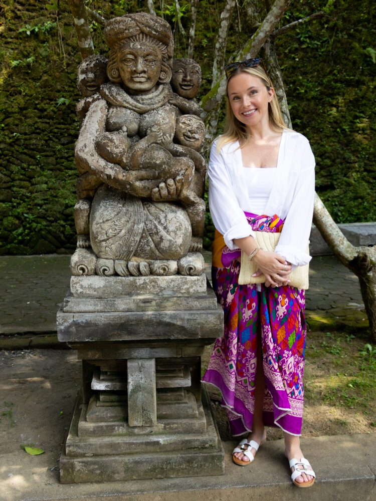Woman in temple attire standing beside an intricately carved stone statue at Pura Tirta Empul temple in Bali.