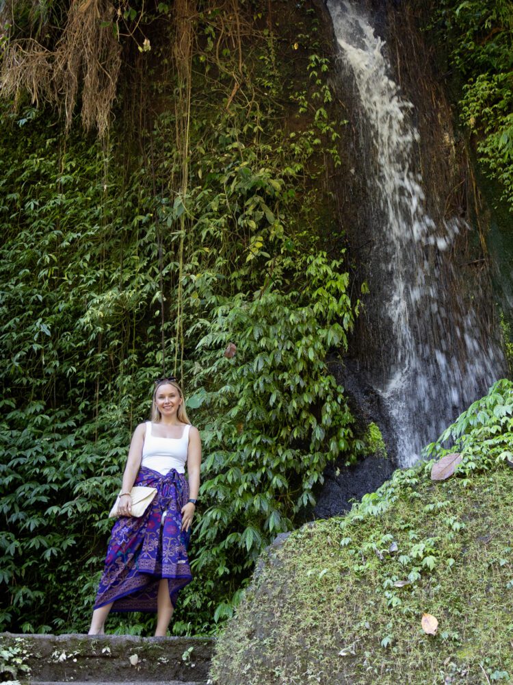 Woman posing near a lush, green-covered waterfall at Pura Gunung Kawi temple complex in Bali.