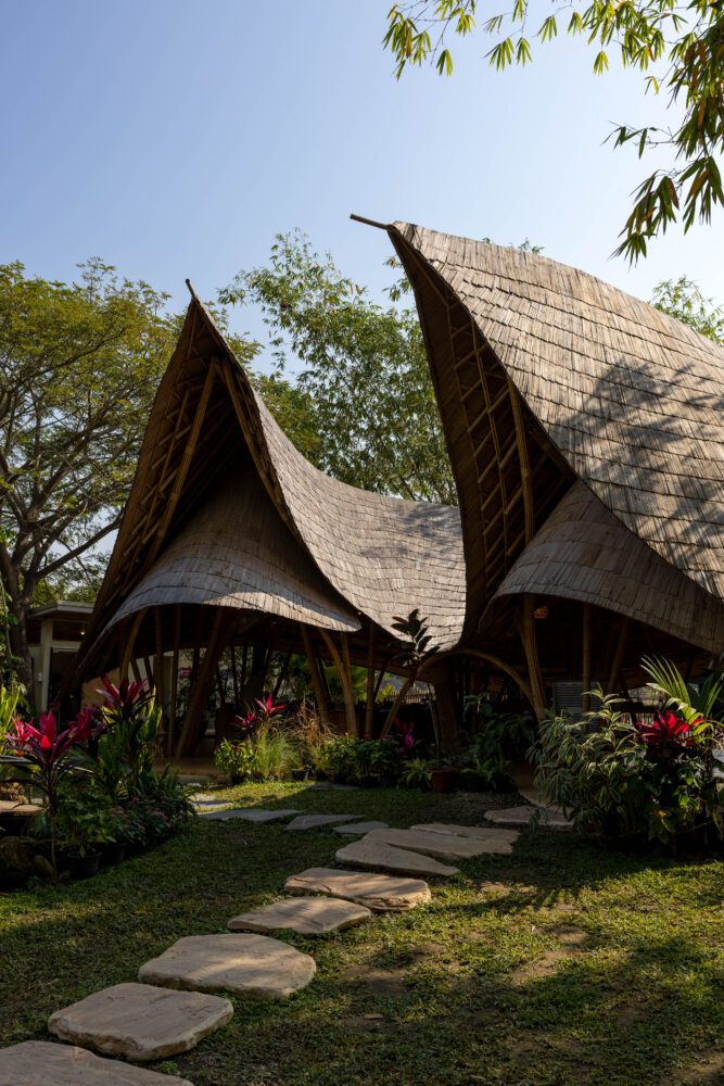 Bamboo-roofed pavilion at Zabb E Lee Cooking School in Chiang Mai, Thailand, surrounded by tropical plants and casting soft shadows in the afternoon sun.