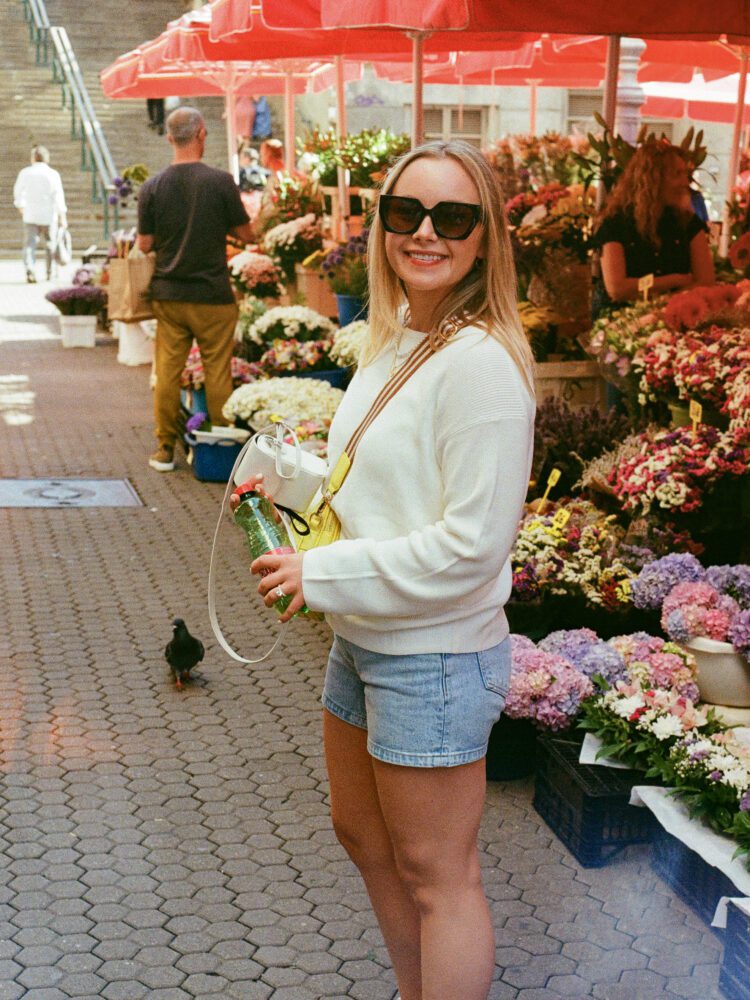 Woman smiling at Zagreb's Dolac Market in Croatia, surrounded by flower stalls featuring vibrant bouquets and red market umbrellas on a sunny day—capturing the charm of local European street markets.