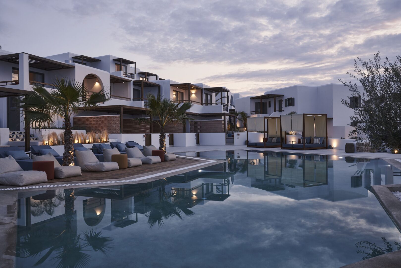 Chic poolside setting at dusk with cabanas, palm trees, and Cycladic-style suites reflecting in the water.