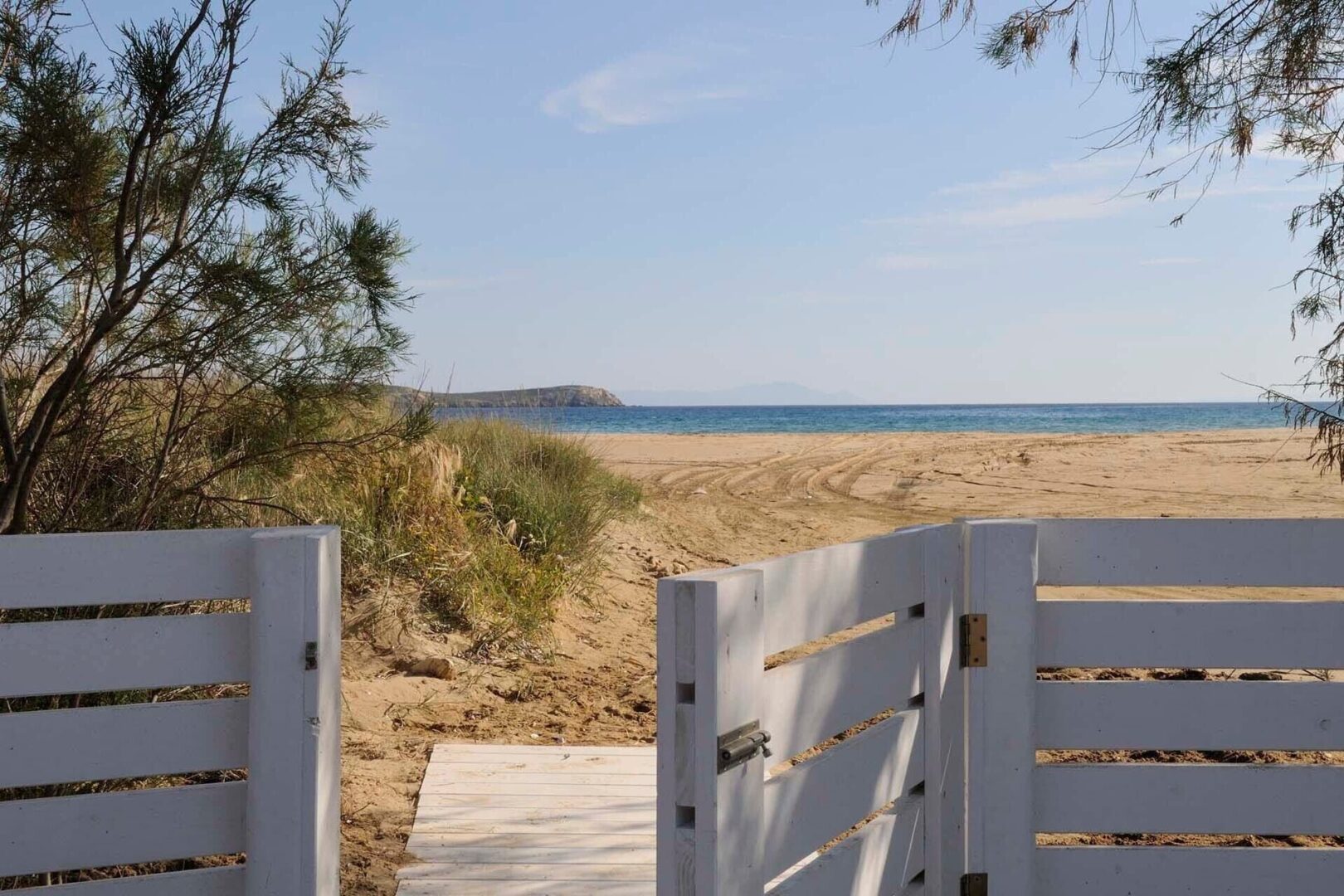 View of Chrissi Akti Golden Beach Paros with sandy shore, blue sea, and a white gate opening to the path.