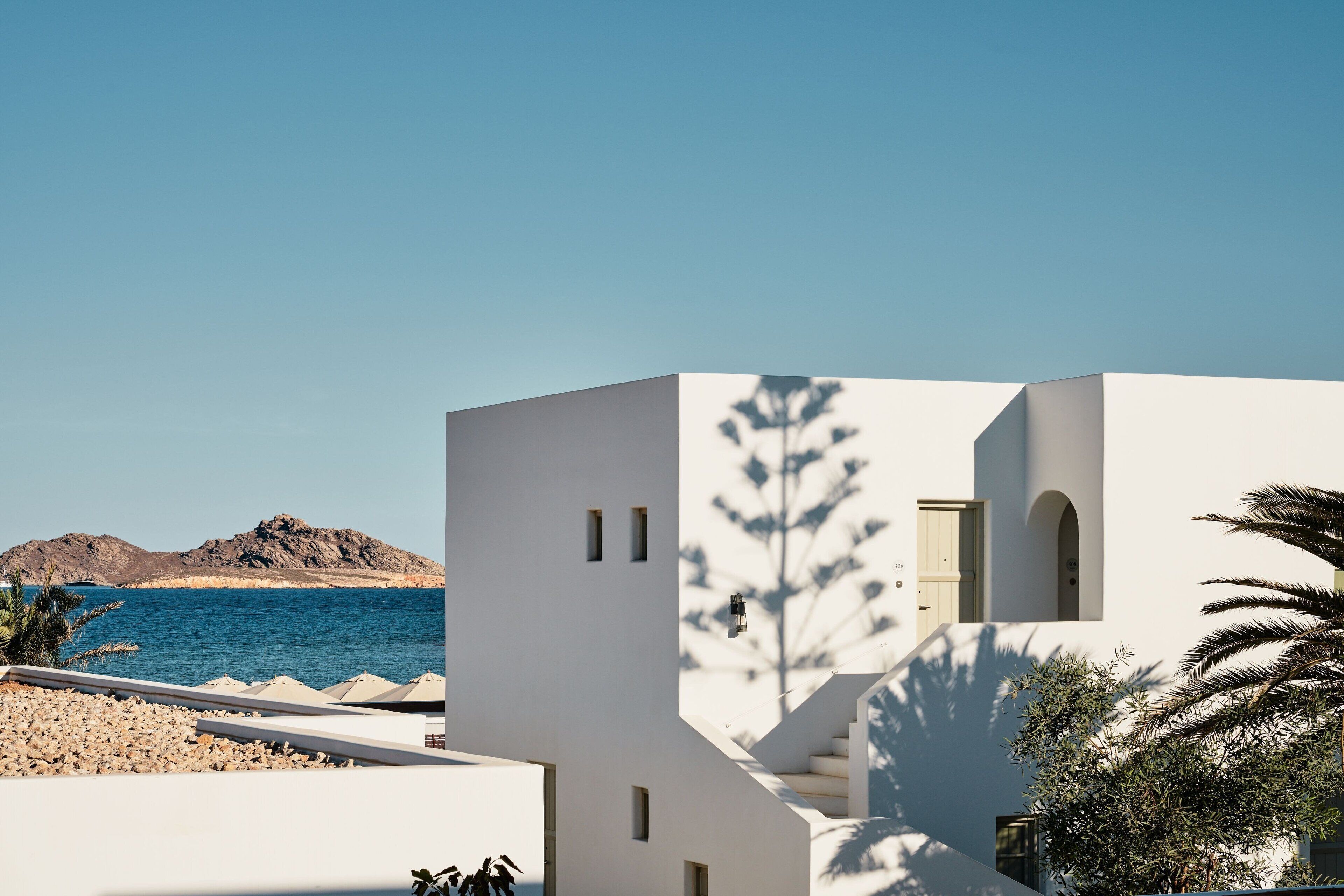 Modern Cycladic architecture with whitewashed walls and sea views framed by palm trees and rocky islets under a clear blue sky.