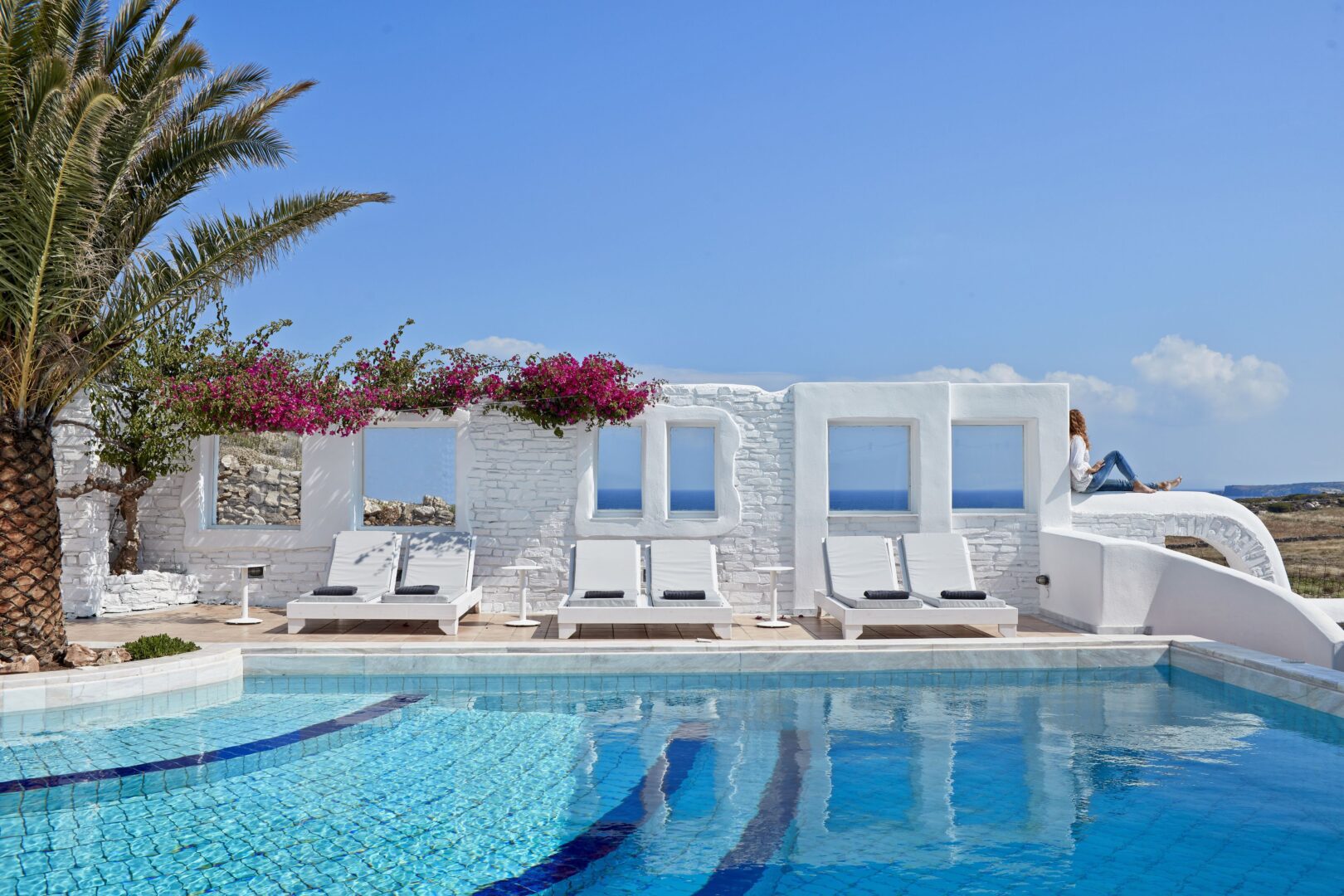 Poolside area at Mr & Mrs White Hotel Paros with palm trees, bougainvillea, and white loungers against a clear blue sky.