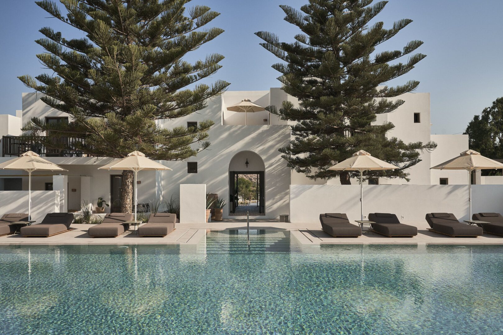 Serene pool area with loungers, tall pines, and minimalist white buildings reflecting Paros island’s signature style.