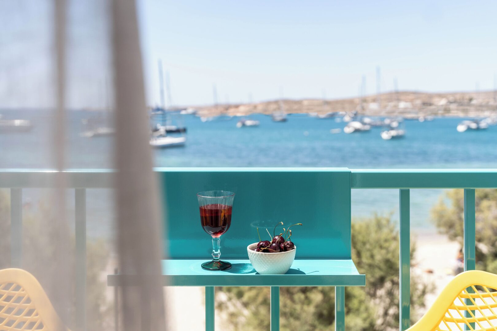 Junior suite balcony at Play Paros Hotel featuring colorful furniture, a glass of wine, and sea views dotted with sailboats.