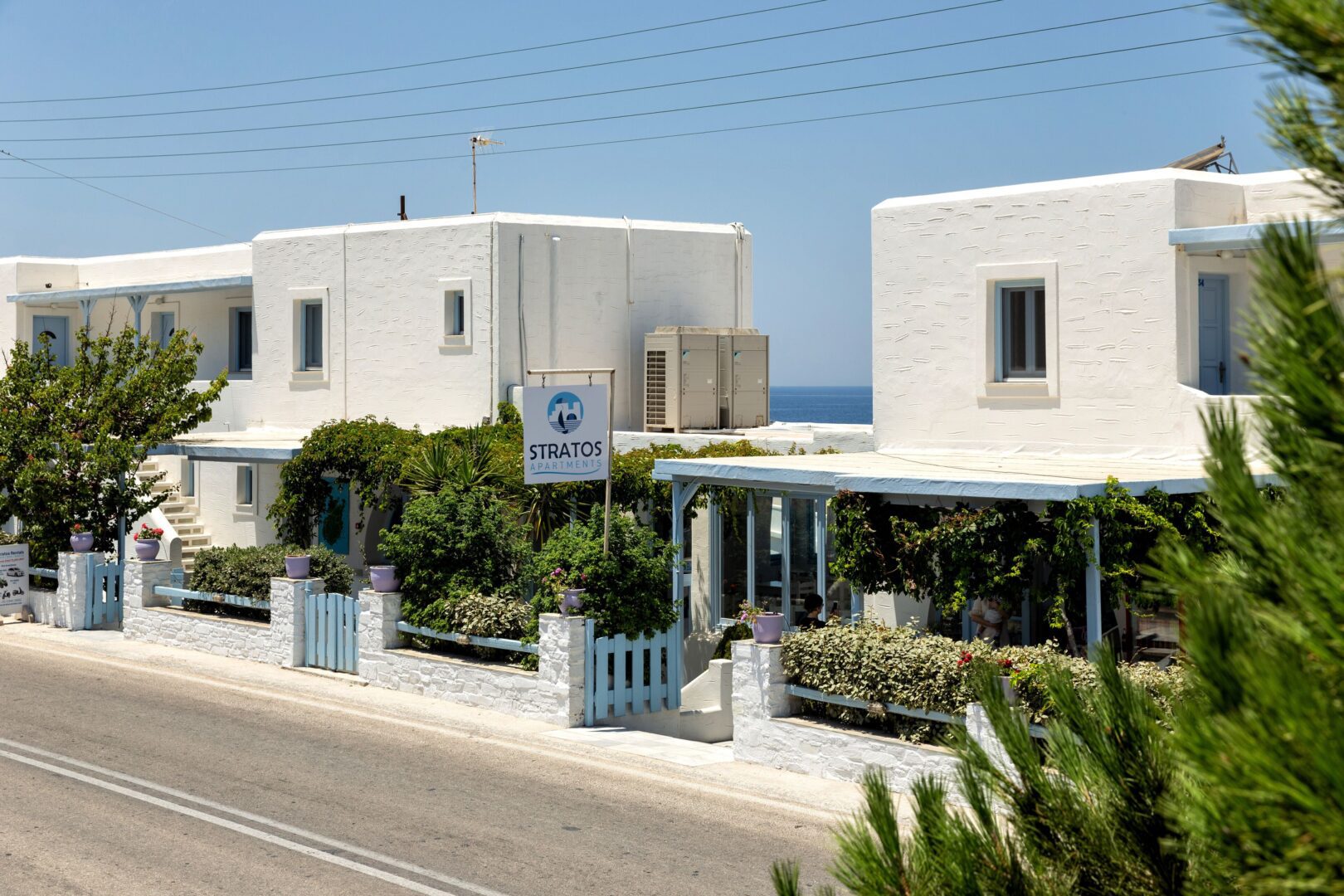 Exterior view of Stratos Apartments & Studios Paros with whitewashed buildings, blue accents, and garden greenery near the sea.