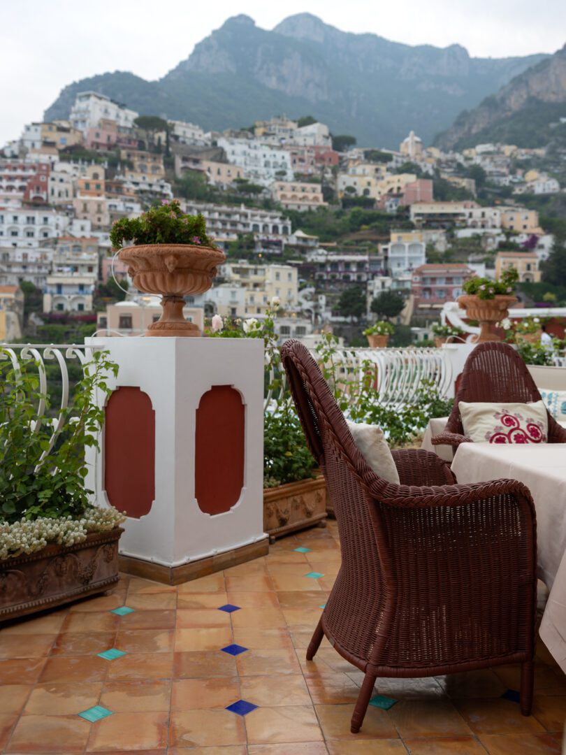 Wicker chairs and tiled terrace at Aldo’s Cocktail Bar, with panoramic views of Positano’s hillside buildings at Le Sirenuse Hotel.