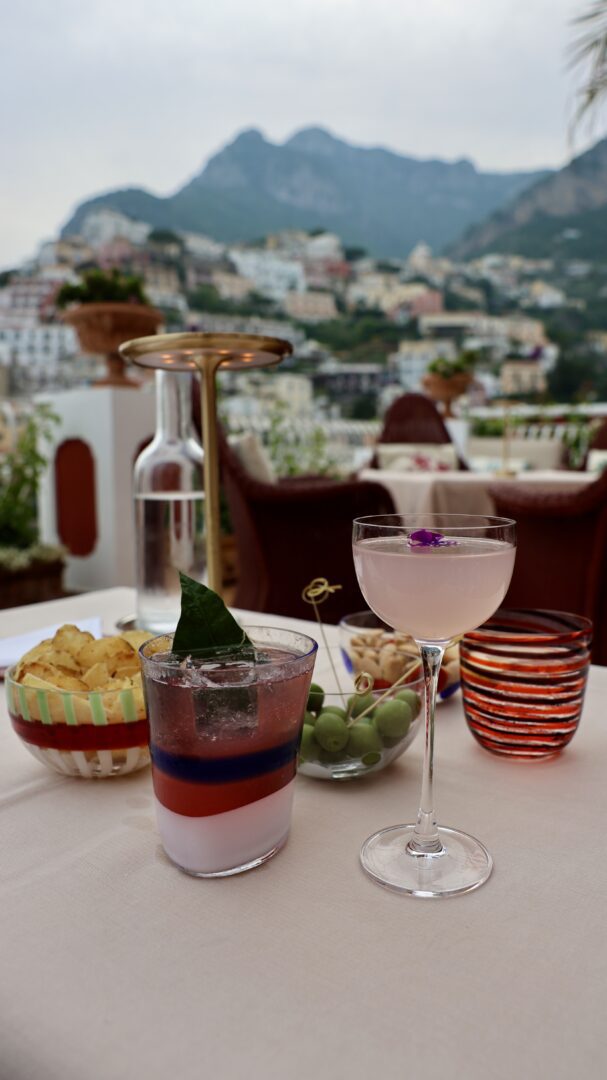 Colorful cocktails and savory snacks are served on a terrace at Aldo’s Cocktail Bar at Le Sirenuse, with Positano’s hillside buildings blurred in the background.