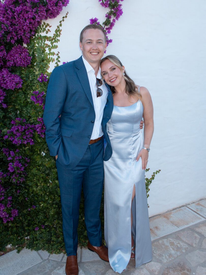 Couple dressed in formal wedding guest attire posing beside vibrant bougainvillea in southern Italy.