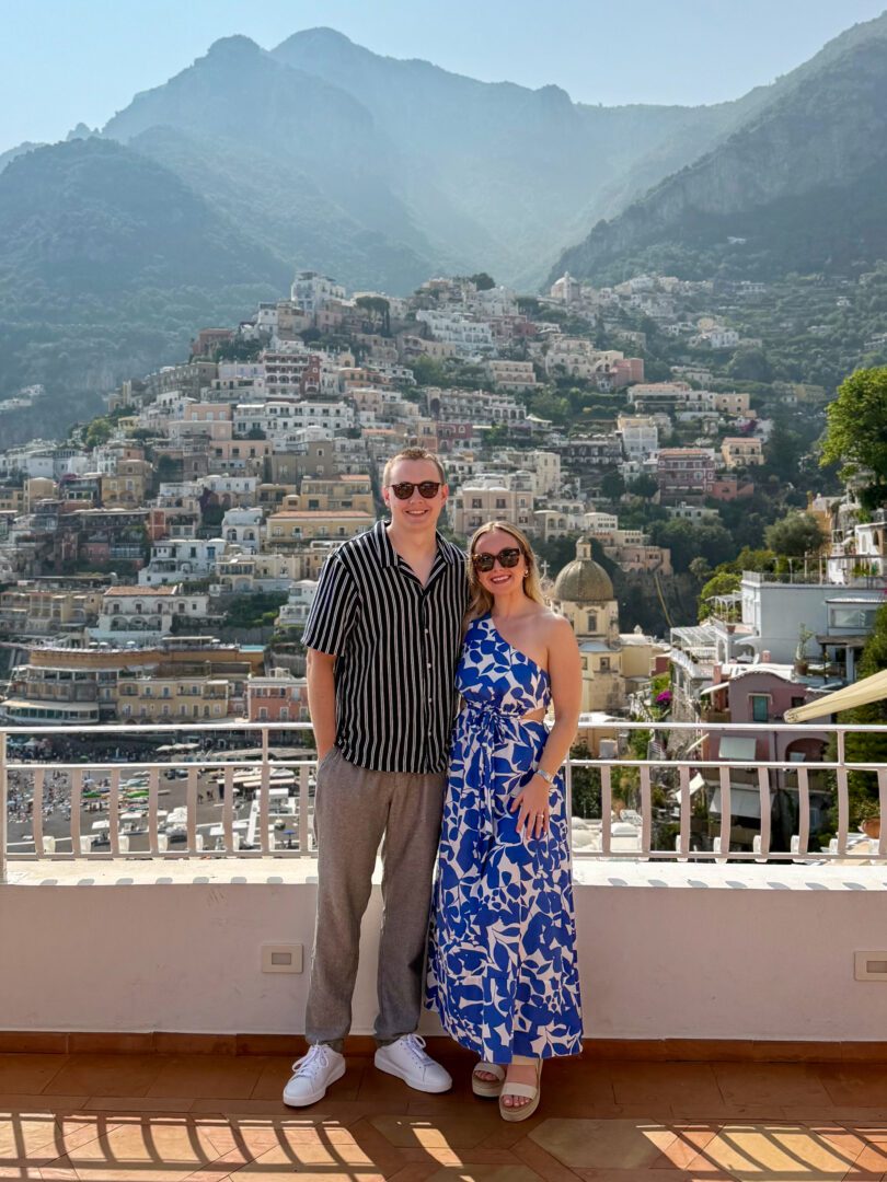 Couple posing on the terrace of Hotel Miramare in Positano, with sun-drenched views of the colorful hillside and the dome of Santa Maria Assunta in the background.