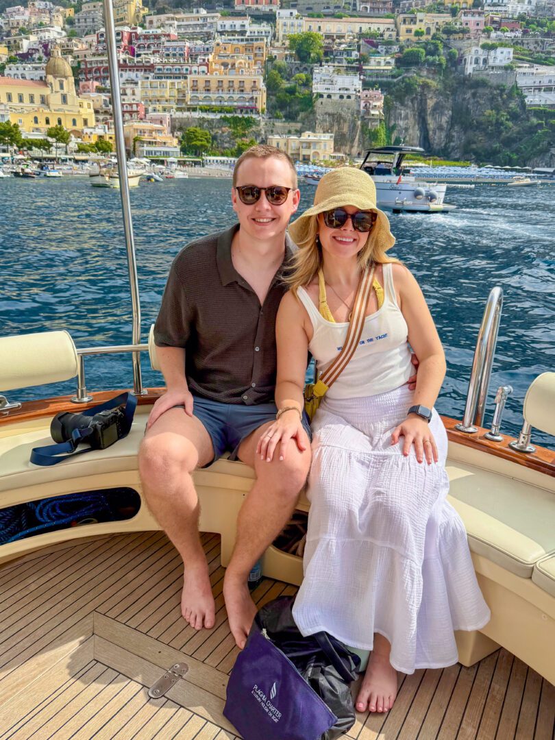 Couple seated on a traditional Italian gozzo boat, cruising away from Positano with the colorful cliffside village in view behind them.