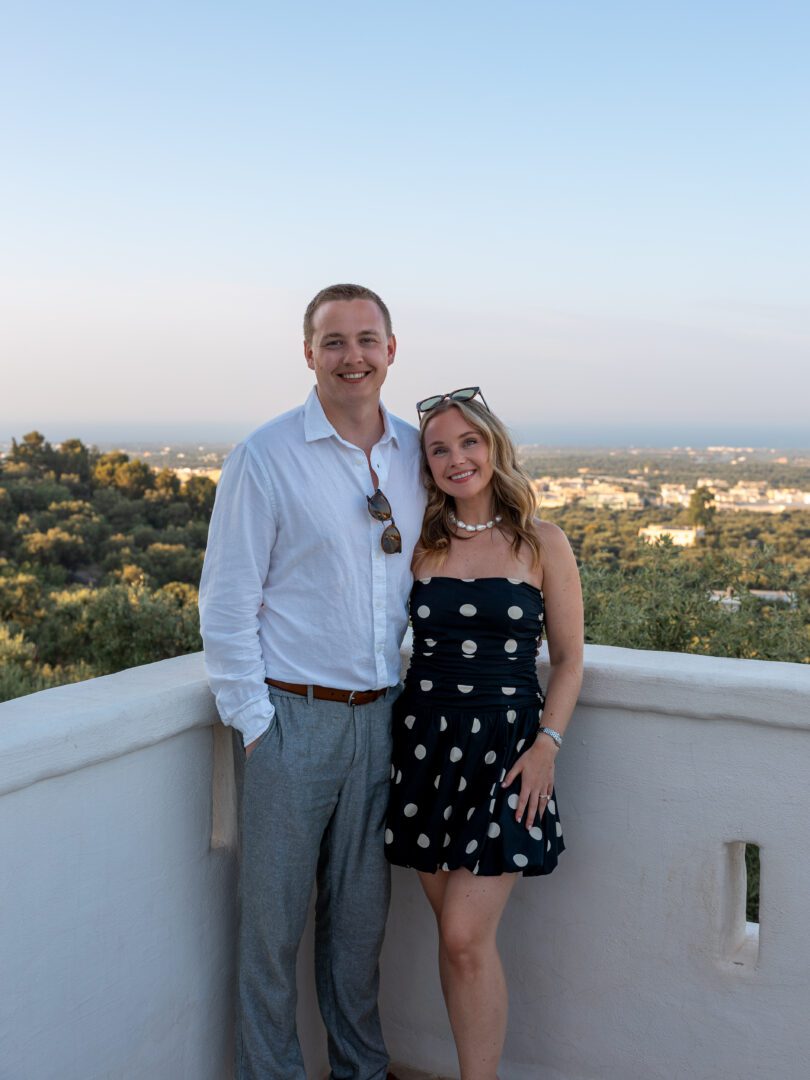 Couple smiling on a whitewashed terrace at Masseria Borgo Ritella in Puglia, with golden hour views of olive groves and countryside in the background.