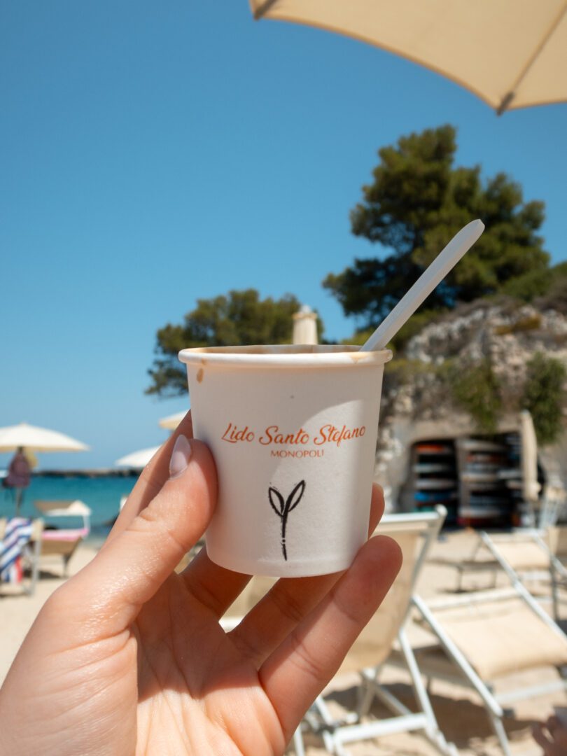Hand holding a crema di caffè cup at Lido Santo Stefano beach club in Monopoli, with umbrellas and sea in the background.