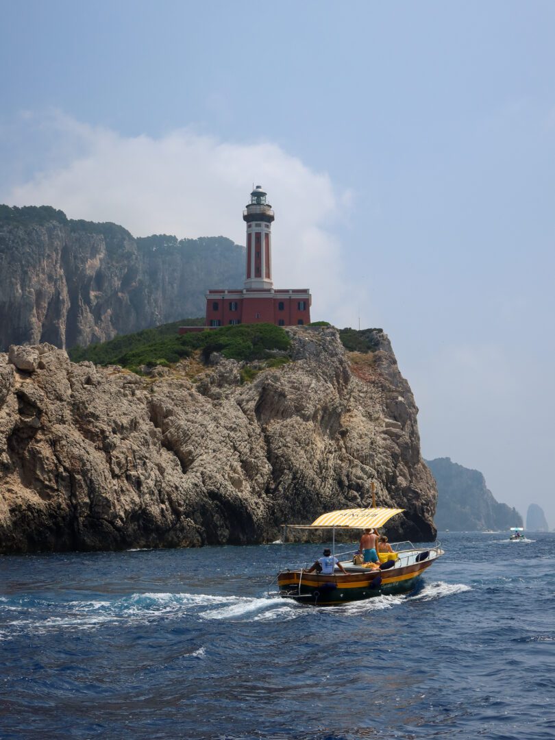 Traditional Italian boat cruising past the Faro di Punta Carena lighthouse on Capri, perched dramatically atop rocky cliffs along the coastline.