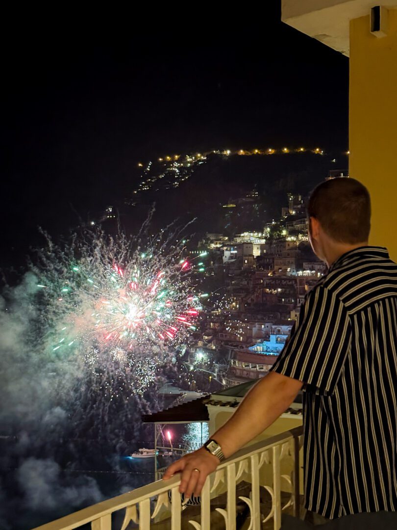 Man watching a vibrant fireworks display over Positano from the balcony of Hotel Miramare, with the hillside aglow against the night sky.