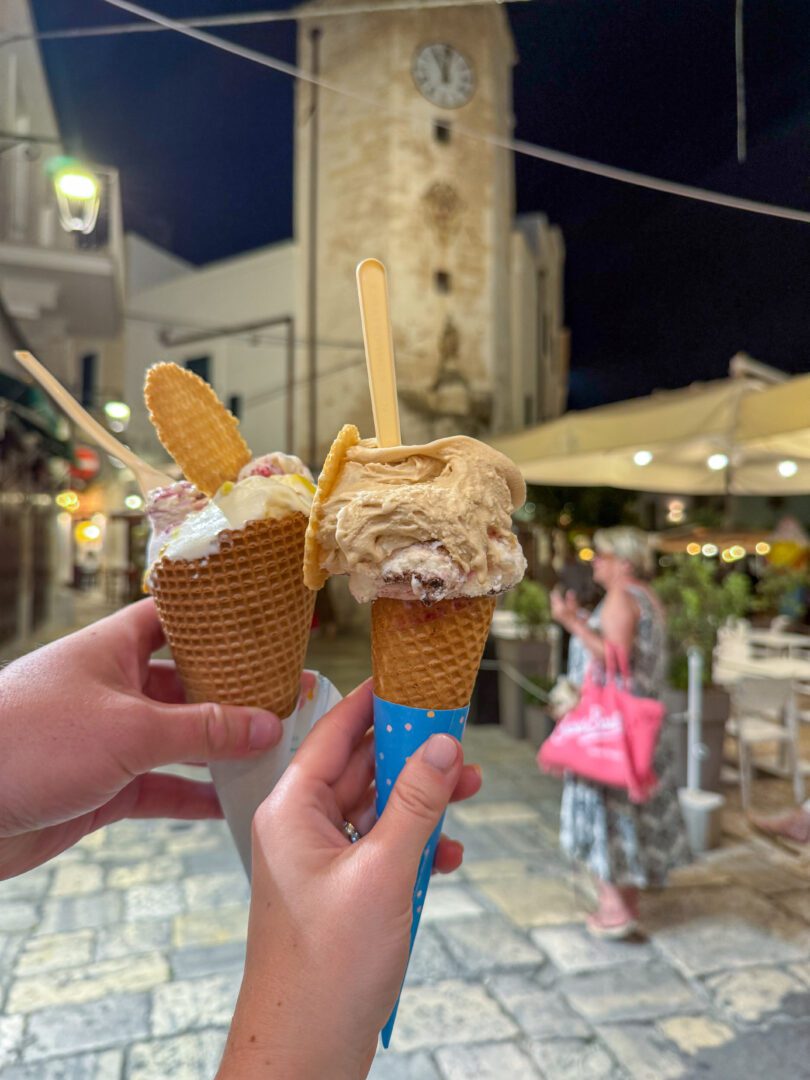 Two gelato cones held up at night in front of Bella Blu Gelateria and a historic clocktower in Monopoli, Italy.