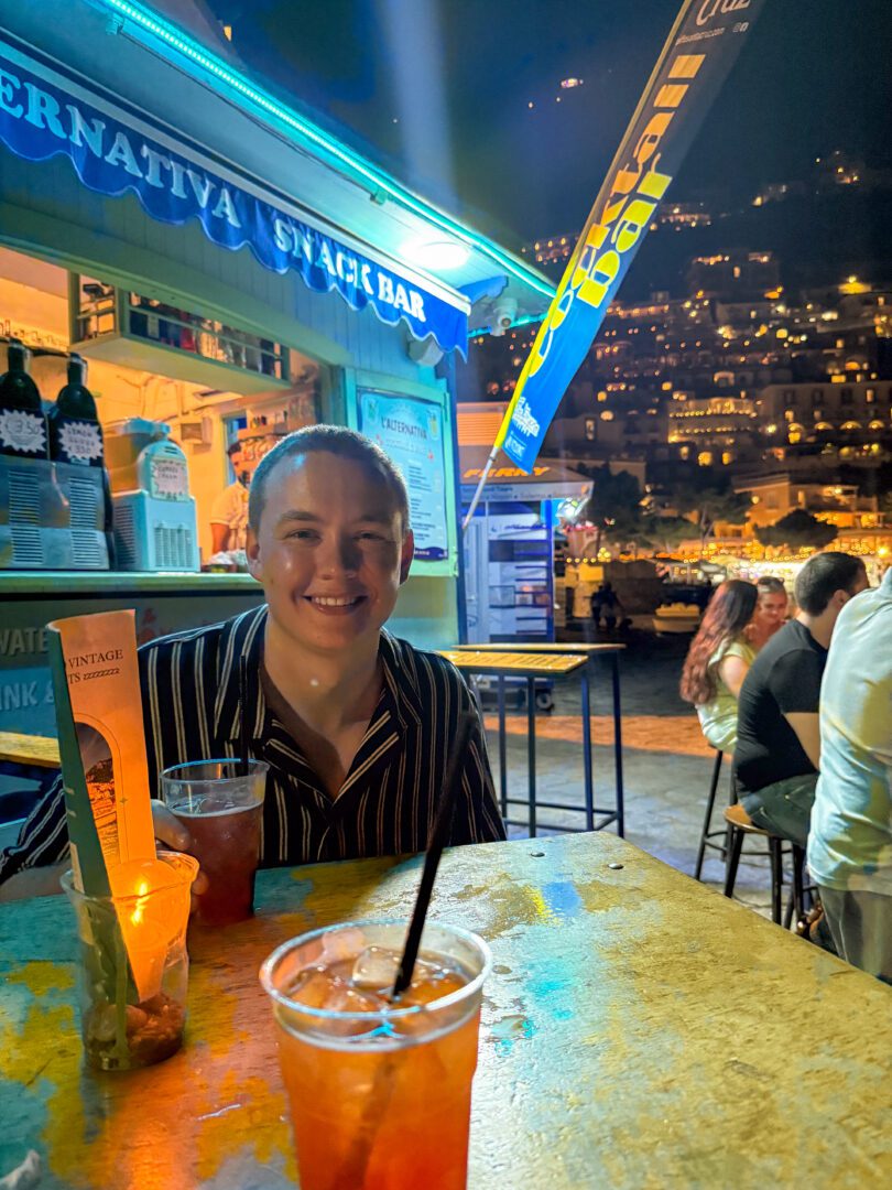 Man enjoying cocktails at L’Alternativa Snack Bar in Positano, with colorful lighting and the hillside town aglow in the background.