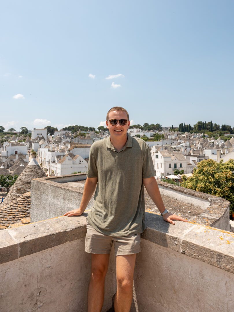 Man smiling at a scenic viewpoint overlooking the traditional trulli houses in Alberobello, Italy.