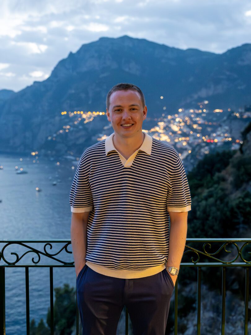 Man standing in front of a railing at Il San Pietro di Positano at dusk, with the Amalfi Coast glowing behind him and lights twinkling along the cliffside.