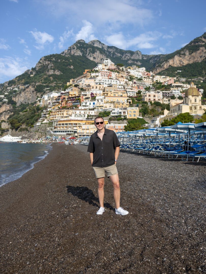 Man stands on Positano’s black pebble beach with a backdrop of colorful cliffside buildings and bright blue skies.