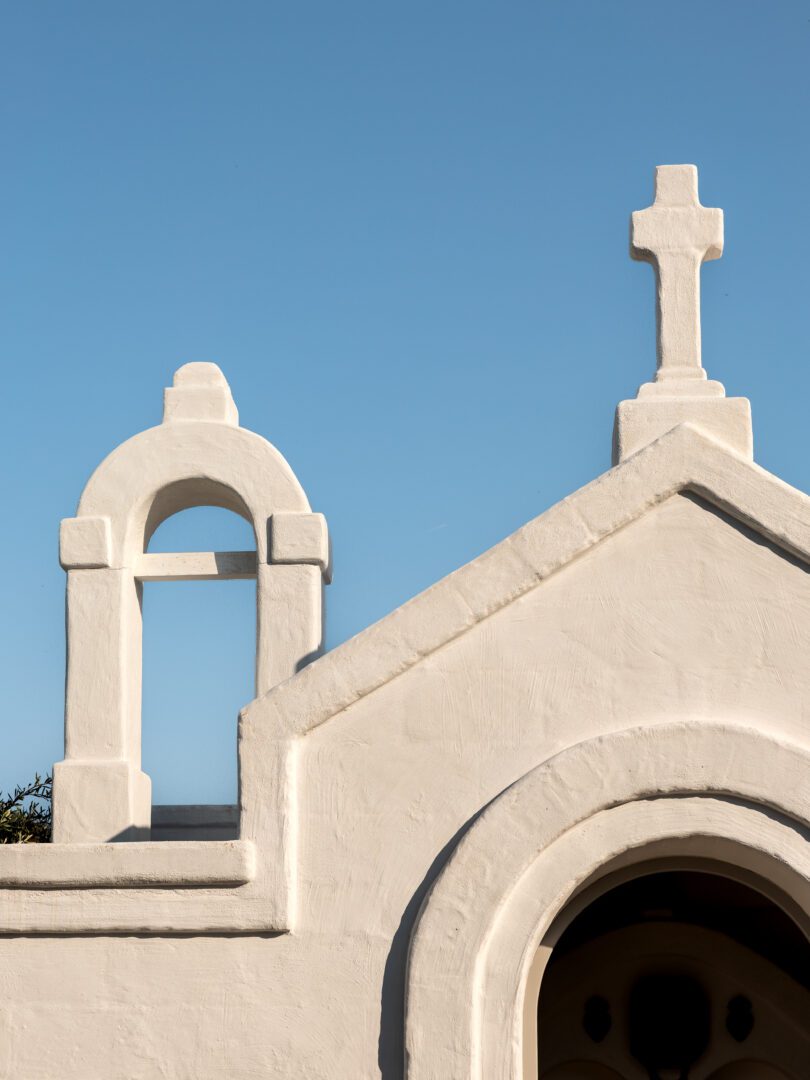 Whitewashed chapel facade with a bell arch and cross at Masseria Borgo Ritella under a clear blue sky in Puglia, Italy.