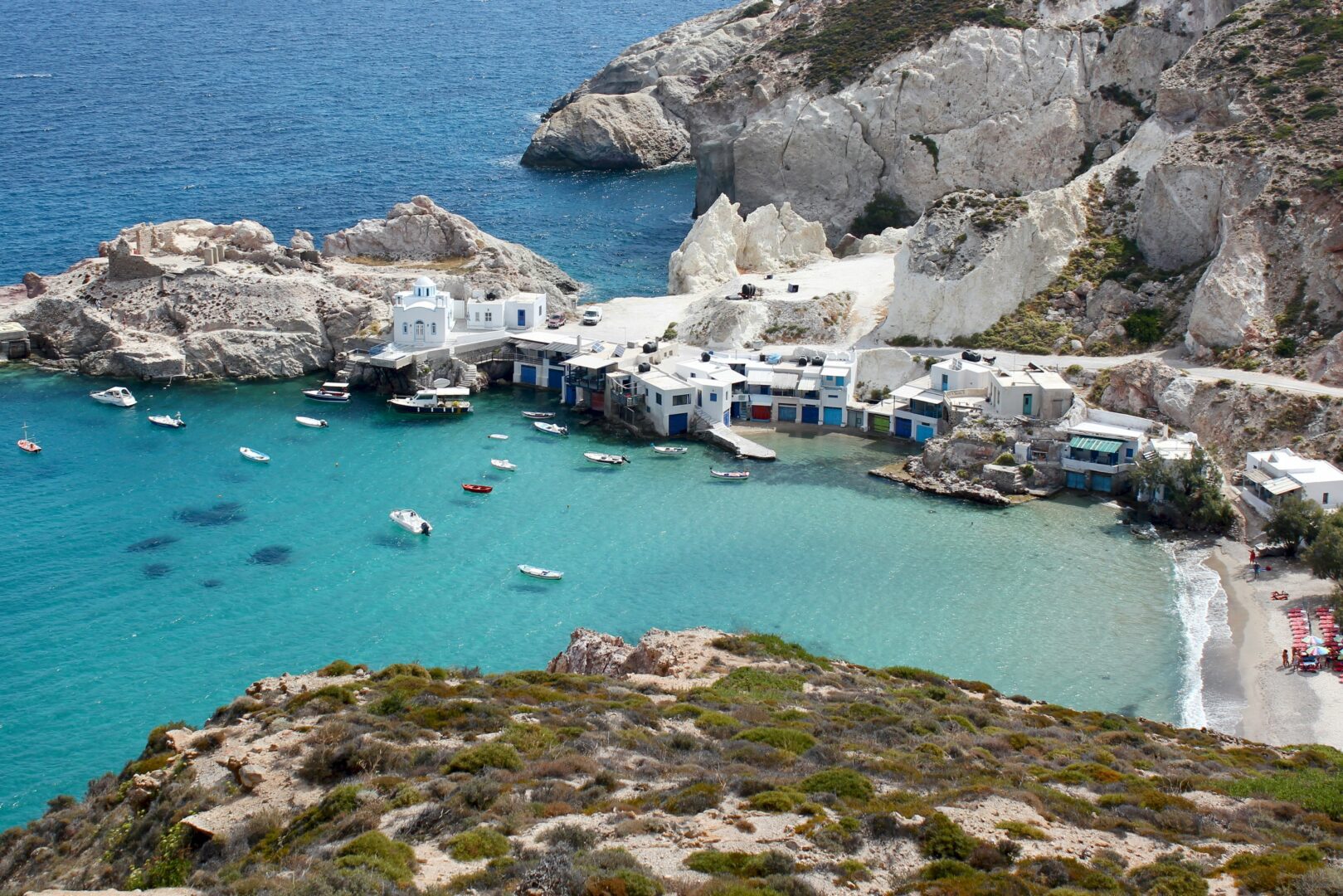Scenic aerial view of Firopotamos's traditional syrmata boathouses along a turquoise bay, framed by rocky cliffs and a whitewashed seaside church.