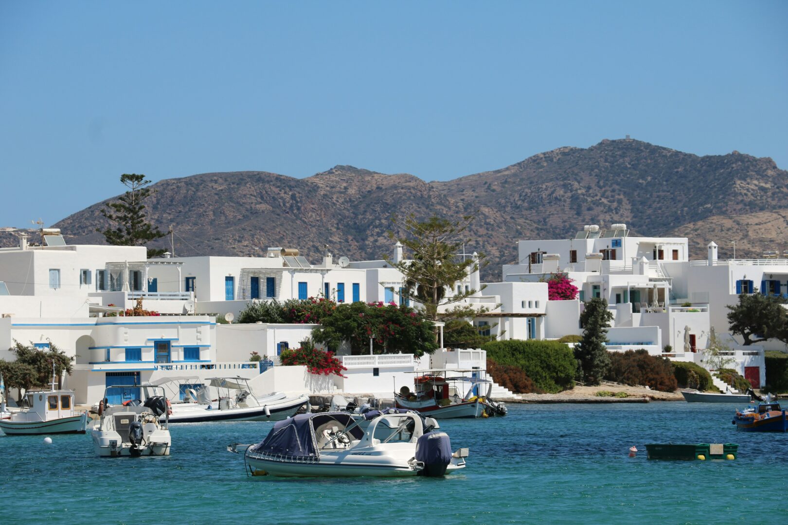 Seaside view of Pollonia, Milos featuring whitewashed Cycladic homes, vibrant bougainvillea, and small fishing boats anchored in the calm turquoise bay.